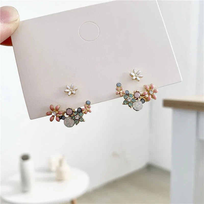 Pair of floral earrings held up against a white card with a blurred indoor background.